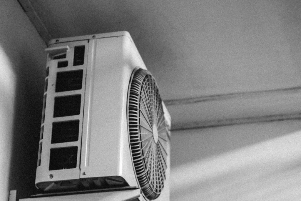 壁の高い位置に金具で取り付けられた、エアコンの室外機を下から見上げた白黒の写真。側面の吸気口と、丸い送風口の格子が見える