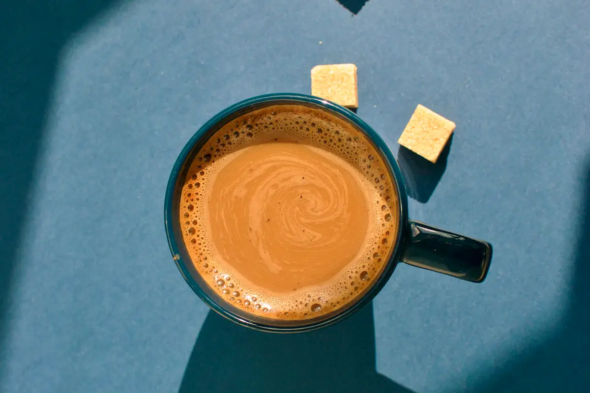 青い背景の上に置いた、ミルク入りのコーヒーのマグカップと角砂糖を上から見た写真