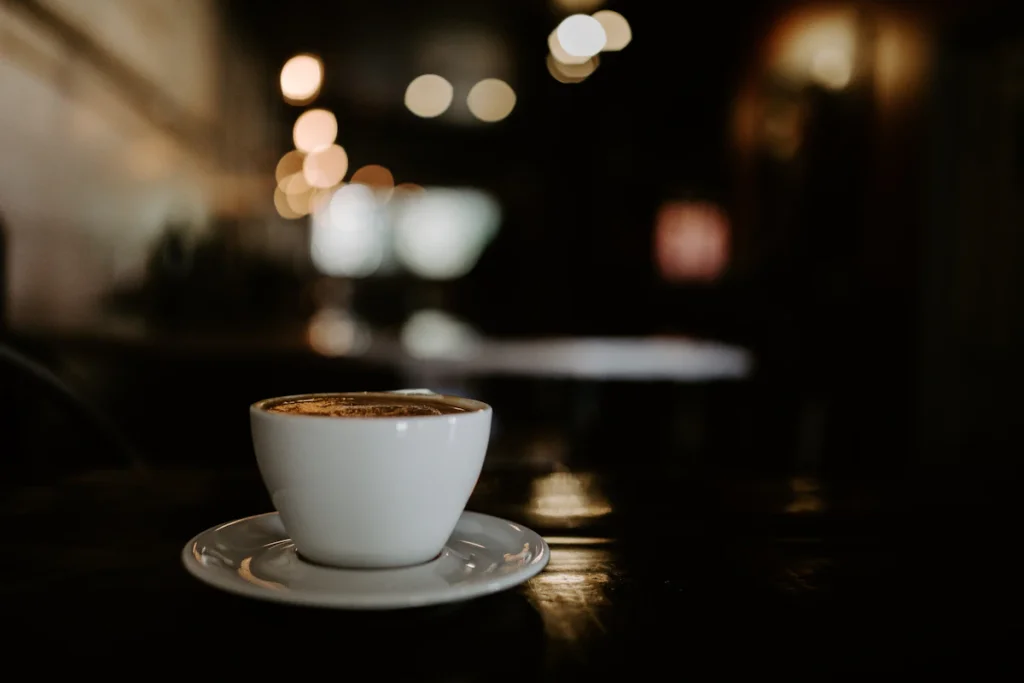 明かりがぼやけて見える暗い室内のテーブルに置いた、受け皿にのせた白いカップのコーヒー