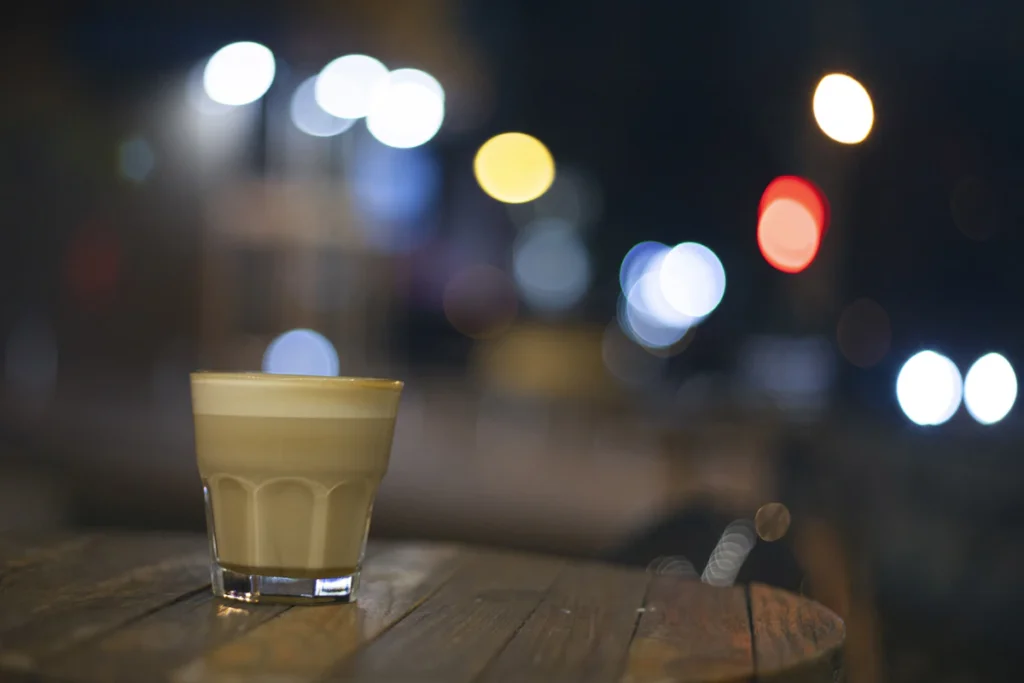 明かりがぼやけて見える暗い背景の前の、木のテーブルに置いた小さなグラス。薄い茶色のコーヒーの上に、泡の層がある