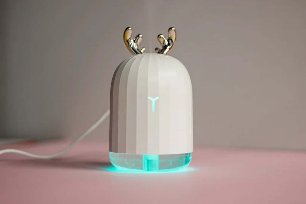 ピンク色の台の上に置いた、白い小型の加湿器。上に金色の角の飾りが付き、下の部分が水色に光っている