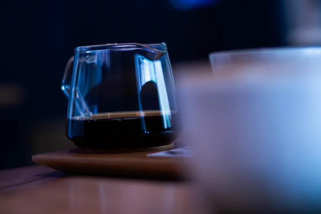 木の皿にのせた、コーヒーの入ったガラスのサーバー。青い光の中で、手前のカップはぼやけている