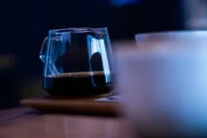木の皿にのせた、コーヒーの入ったガラスのサーバー。青い光の中で、手前のカップはぼやけている